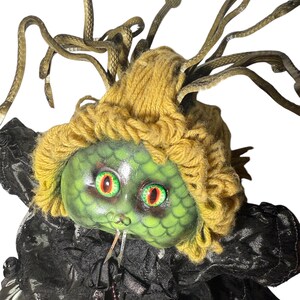 Pode incluir: Uma boneca Medusa com rosto verde, cabelo de fio amarelo e cabelo de cobra bronze. A boneca tem olhos vermelhos e verdes e usa uma roupa preta com babados. As cobras s&atilde;o posicionadas ao redor da cabe&ccedil;a.