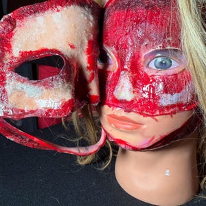 Puede incluir: Una máscara de Halloween y una cabeza de maniquí con una apariencia sangrienta y sangrienta. La máscara es blanquecina con detalles en rojo, mientras que la cabeza está cubierta de pintura roja, simulando un efecto macabro. El maniquí tiene cabello rubio y ojos azules.
