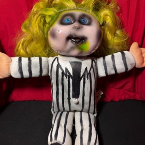 Pu&ograve; includere: Bambola fatta a mano con viso dipinto, occhi blu e capelli verdi e gialli. La bambola indossa un abito a righe bianche e nere e ha i piedi neri. Le braccia della bambola sono tese.