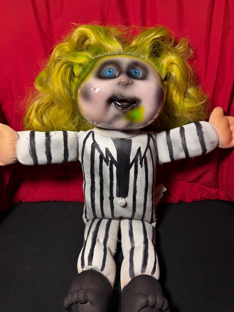 Pu&ograve; includere: Bambola fatta a mano con viso dipinto, occhi blu e capelli verdi e gialli. La bambola indossa un abito a righe bianche e nere e ha i piedi neri. Le braccia della bambola sono tese.