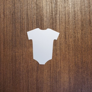 May include: White paper cutout of a baby onesie on a brown wood background.