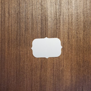 May include: A white blank label with a decorative border, on a brown wood background.