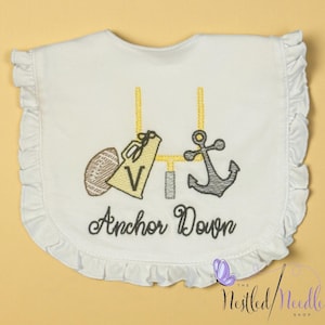 May include: White baby bib with a ruffled edge, embroidered with a football, megaphone, anchor, and goalpost. The text "Anchor Down" is embroidered below the images. The bib is on a yellow background.