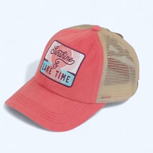 May include: A coral-colored baseball cap with a mesh back and a rectangular patch. The patch features the words "Sunshine & Lake Time" with a sun graphic. The cap has a curved brim and a tan mesh back.