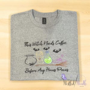 May include: A gray t-shirt with the embroidered phrase "This Witch Needs Coffee Before Any Hocus Pocus." The design includes a cauldron, coffee cup, potion bottle, bats, and a broom. The shirt is folded neatly.