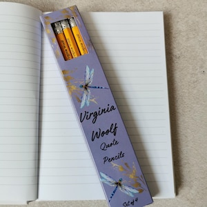 May include: A lavender box containing four pencils, labelled "Virginia Woolf Quote Pencils." The box is decorated with a dragonfly design and gold accents. The pencils are yellow with silver erasers.