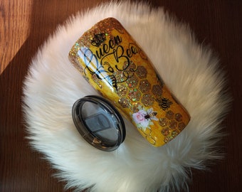 May include: A yellow and gold tumbler with a black and white design that says "Queen Bee". The tumbler has a honeycomb pattern and glitter accents. It is sitting on a white fluffy surface.