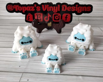 May include: Three white Yeti figurines with blue faces and arms. The Yeti are sitting on a white wooden surface. The Yeti are approximately 4 inches tall.
