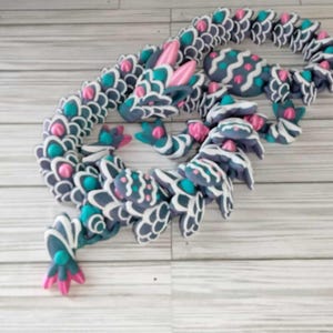 Puede incluir: Un dragón enrollado de arcilla polimérica, de color gris, blanco y rosa. El dragón tiene un diseño detallado con escamas y una superficie texturizada.