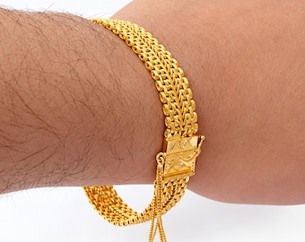 May include: A gold bracelet featuring a multi-strand woven chain design with a rectangular box clasp. The clasp is decorated with an engraved floral pattern and includes a small, dangling chain loop attached to the side of the closure mechanism.