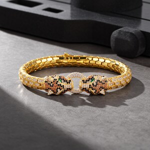 May include: A gold-toned bracelet featuring a honeycomb-patterned band and two leopard head terminals. The leopard heads are encrusted with small clear, black, and orange stones, with green stone eyes, and they hold a circular clear stone ring.