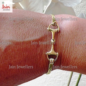 May include: A gold-coloured bracelet with a horse bit design. The bracelet is made of metal and features a curved hook clasp. The jewellery is worn on a wrist.