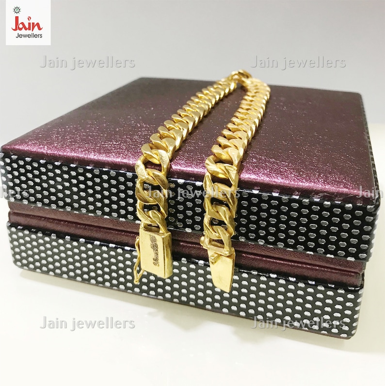 May include: Two gold chains with a curb link design are displayed on a burgundy and black patterned box. The box has the text "Jain Jewellers" printed on it.