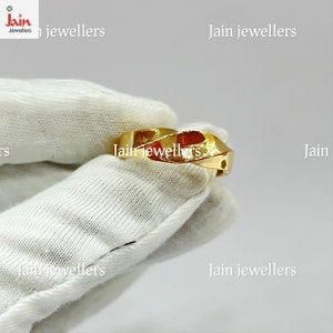 Puede incluir: Un anillo de oro con un dise&ntilde;o retorcido. El anillo est&aacute; hecho de un metal amarillo brillante y tiene un patr&oacute;n geom&eacute;trico. El anillo se muestra sobre una superficie blanca. El texto "Jain jewellers" es visible.