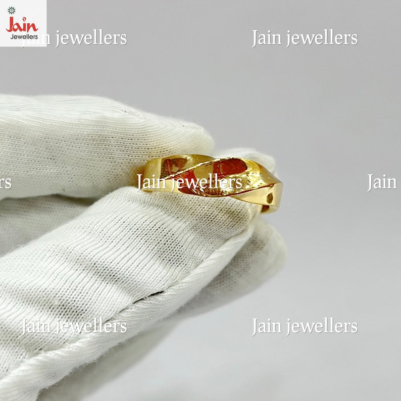 Puede incluir: Un anillo de oro con un dise&ntilde;o retorcido. El anillo est&aacute; hecho de un metal amarillo brillante y tiene un patr&oacute;n geom&eacute;trico. El anillo se muestra sobre una superficie blanca. El texto "Jain jewellers" es visible.