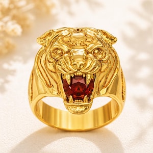 May include: A gold ring with a tiger head design. The tiger's open mouth reveals a red gemstone. The ring has a polished finish, highlighting the detailed craftsmanship. The ring is a statement piece of jewellery.