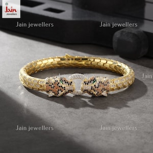 May include: A gold bangle bracelet with two leopard head clasps. The leopard heads are encrusted with clear and black stones, with green accents for the eyes. The bracelet is made of a textured gold metal.
