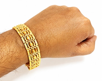 May include: A wide gold-tone bracelet featuring a complex, multi-link chain design with repeating rectangular and circular segments. The metal surface displays a polished finish with intricate etched patterns on the outer rectangular links.