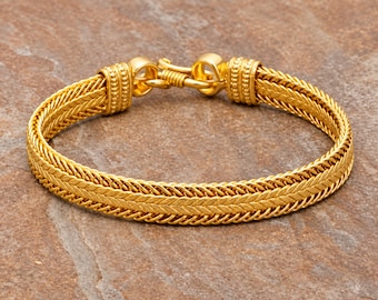 May include: A gold-toned bracelet featuring a woven, herringbone-style chain pattern. The piece is secured with a hook-and-eye clasp mechanism, flanked by two cylindrical end caps decorated with a series of small, raised spherical beads.