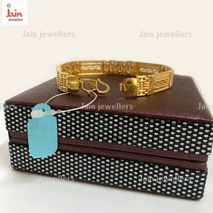 Puede incluir: Un brazalete de oro con un dise&ntilde;o geom&eacute;trico. El brazalete est&aacute; sobre una caja marr&oacute;n con un patr&oacute;n blanco y negro. La caja tiene el texto "Jain Jewellers".