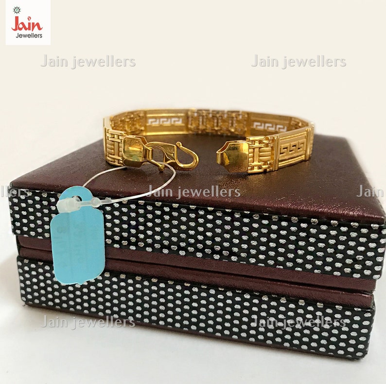 Puede incluir: Un brazalete de oro con un dise&ntilde;o geom&eacute;trico. El brazalete est&aacute; sobre una caja marr&oacute;n con un patr&oacute;n blanco y negro. La caja tiene el texto "Jain Jewellers".