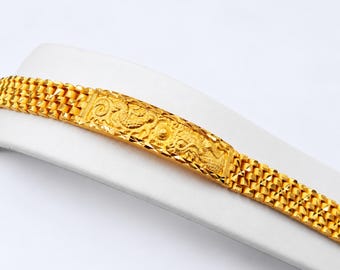 May include: A gold bracelet featuring a central rectangular plaque with an embossed dragon design, flanked by a textured, multi-link chain. The metal has a bright, reflective finish with intricate detailing on the central panel.