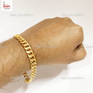 May include: A gold curb chain bracelet with a thick, flat design. The bracelet is shown on a person's wrist.
