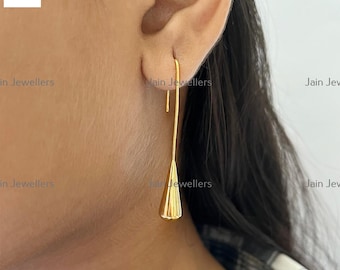 Puede incluir: Un solo pendiente dorado con un gancho largo y delgado y un colgante cónico texturizado. El pendiente se muestra sobre un fondo borroso, resaltando su elegante diseño. Una pieza de joyería sencilla pero elegante.