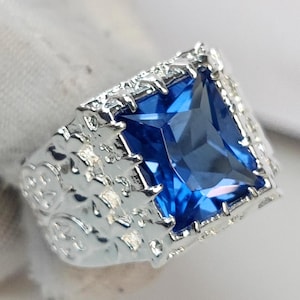 May include: A silver ring featuring a large, square-shaped blue gemstone set in an intricate, ornate design.