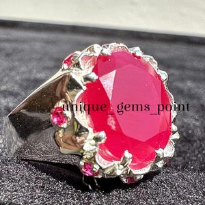 May include: A silver ring featuring a large, faceted, oval-shaped, bright pink gemstone set in a crown-like setting with smaller pink gemstones around the perimeter. The text "unique_gems_point" is visible on the ring.