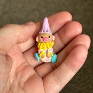 May include: A small, hand-painted clay figurine of a clown with a pink hat, yellow and pink polka dot outfit, and blue shoes.