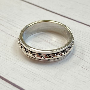 May include: A silver ring with a braided design. The ring is on a white wooden surface.