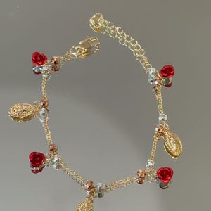 May include: A gold-tone chain bracelet featuring alternating small red rose charms and oval-shaped religious medals. The bracelet is accented with tiny metallic beads in silver and copper tones, finished with a lobster claw clasp and an extender chain.