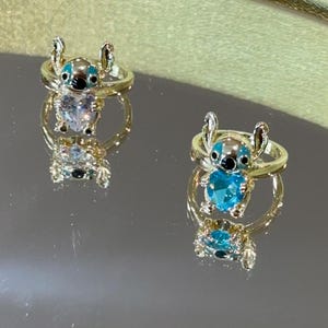 Puede incluir: Dos anillos dorados con un dise&ntilde;o del personaje Stitch. Cada anillo presenta una figura de Stitch con detalles azules y negros y una piedra preciosa azul en forma de coraz&oacute;n. Los anillos se muestran sobre una superficie reflectante.