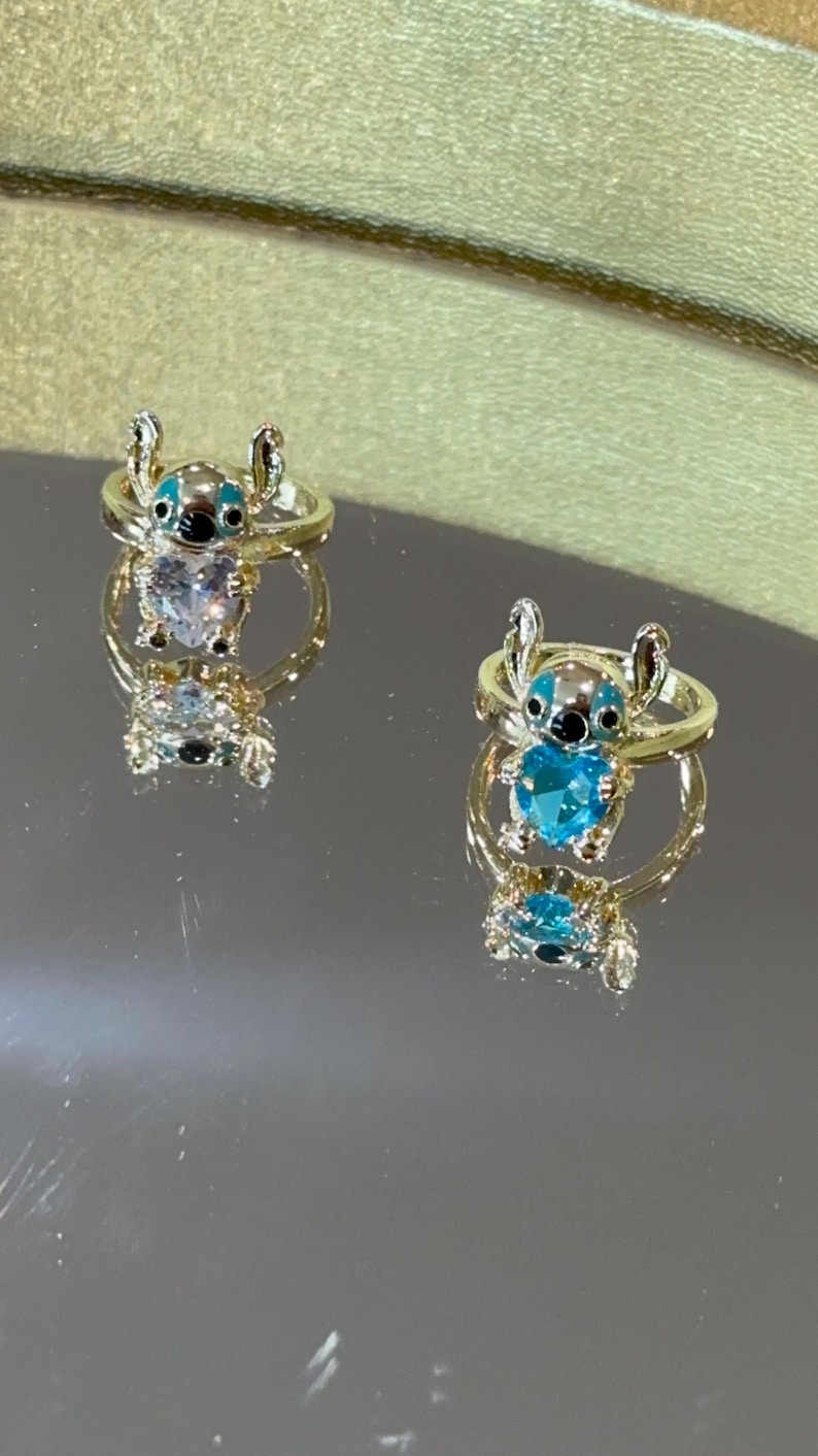 Puede incluir: Dos anillos dorados con un dise&ntilde;o del personaje Stitch. Cada anillo presenta una figura de Stitch con detalles azules y negros y una piedra preciosa azul en forma de coraz&oacute;n. Los anillos se muestran sobre una superficie reflectante.