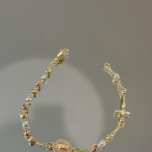 Könnte beinhalten: Goldfarbenes Rosenkranzarmband mit einem Kreuzanhänger und einem Medaillon mit einer religiösen Figur. Das Armband hat eine Mischung aus goldfarbenen, roségoldfarbenen und silberfarbenen Perlen. Der Verschluss ist ebenfalls goldfarben.