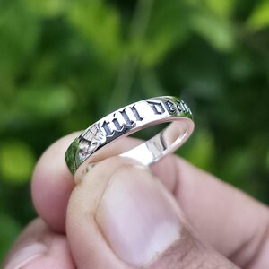 May include: A silver ring with the words "still death" engraved in a gothic font. The ring is held against a blurred green background, highlighting the inscription and the ring's polished surface.
