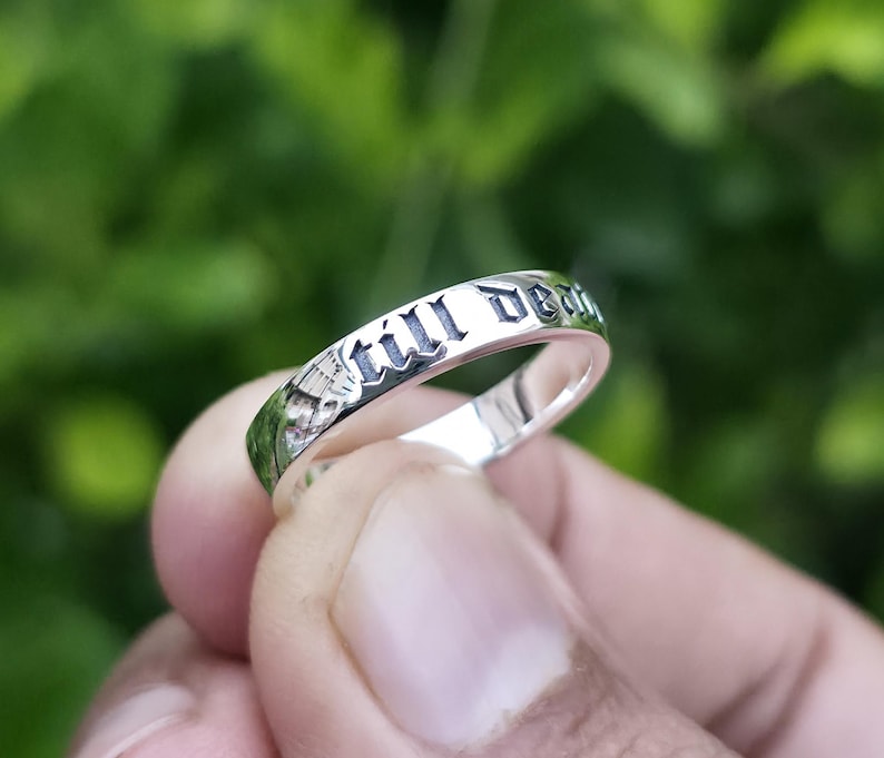 May include: A silver ring with the words "still death" engraved in a gothic font. The ring is held against a blurred green background, highlighting the inscription and the ring's polished surface.