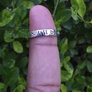 May include: A silver ring with the words "still dead" engraved around the band. The ring is displayed on a finger against a backdrop of green foliage.