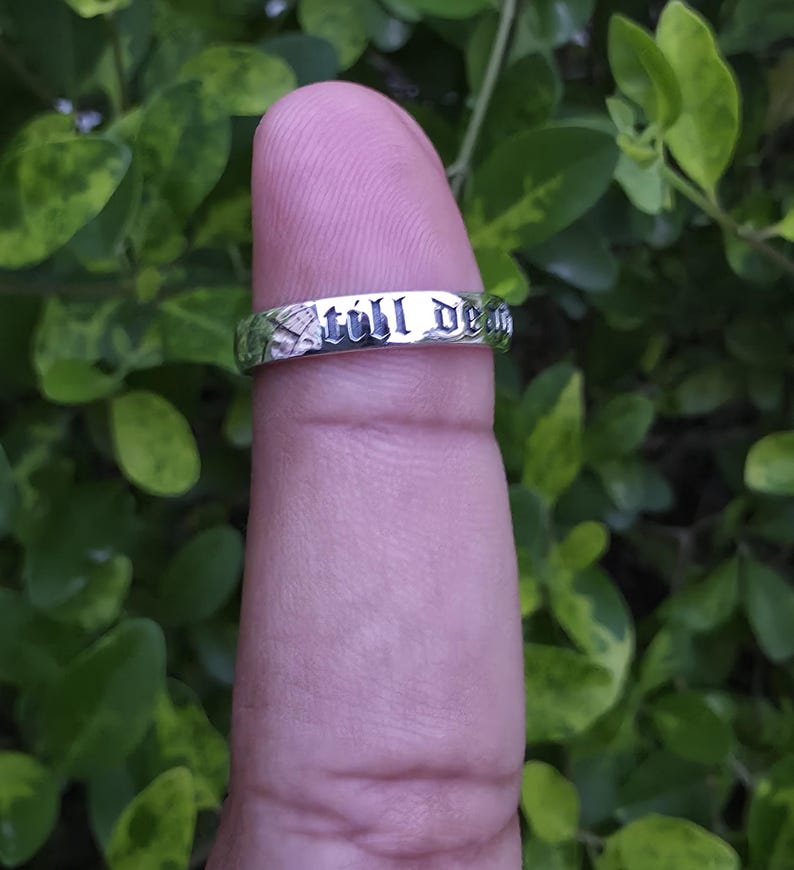 May include: A silver ring with the words "still dead" engraved around the band. The ring is displayed on a finger against a backdrop of green foliage.