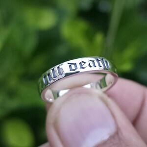 May include: A silver ring with the inscription "till death" in a gothic font. The ring is smooth and shiny, reflecting the light. The background is a blurred green, suggesting an outdoor setting.