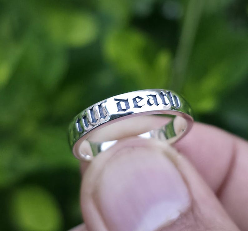 May include: A silver ring with the inscription "till death" in a gothic font. The ring is smooth and shiny, reflecting the light. The background is a blurred green, suggesting an outdoor setting.