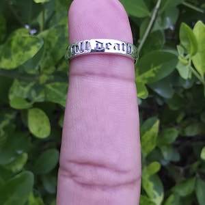 May include: A silver ring with the words "Till Death" engraved in a gothic font. The ring is displayed on a finger against a backdrop of green foliage.
