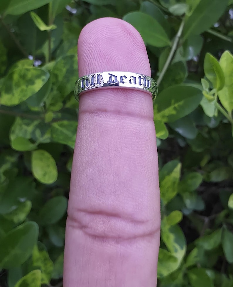 May include: A silver ring with the words "Till Death" engraved in a gothic font. The ring is displayed on a finger against a backdrop of green foliage.