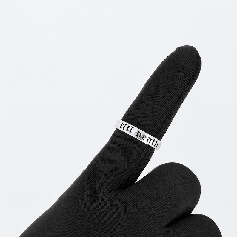 May include: A silver ring with the words "till death" engraved in a gothic font. The ring is displayed on a black surface, creating a contrast that highlights the inscription.
