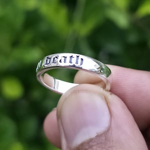 May include: A silver ring with the word "Death" engraved in a gothic font. The ring is held up against a blurred green background, showcasing its polished surface and detailed inscription.