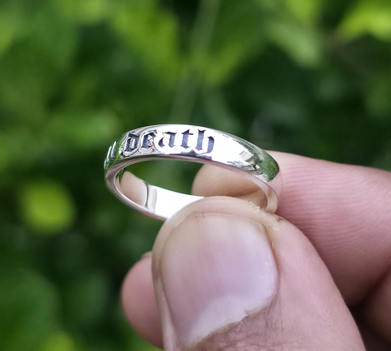 May include: A silver ring with the word "Death" engraved in a gothic font. The ring is held up against a blurred green background, showcasing its polished surface and detailed inscription.