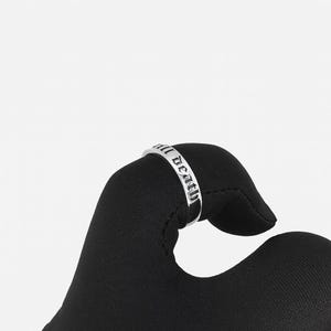 May include: A silver ring with the inscription "all death" in a gothic font. The ring is held against a black background, showcasing the polished metal and the detailed lettering. The ring's design is simple yet bold.