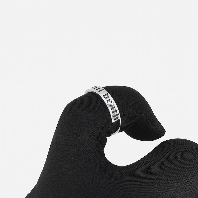May include: A silver ring with the inscription "all death" in a gothic font. The ring is held against a black background, showcasing the polished metal and the detailed lettering. The ring's design is simple yet bold.