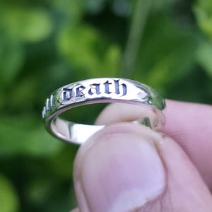 May include: A silver ring with the word "death" engraved in a gothic font. The ring is held in a hand against a blurred green background. The ring has a polished finish.